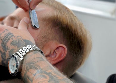 Close-up of shaving at Barber Boy Studio, Horsham, England, GB. Tattooed hand skillfully holds straight razor.