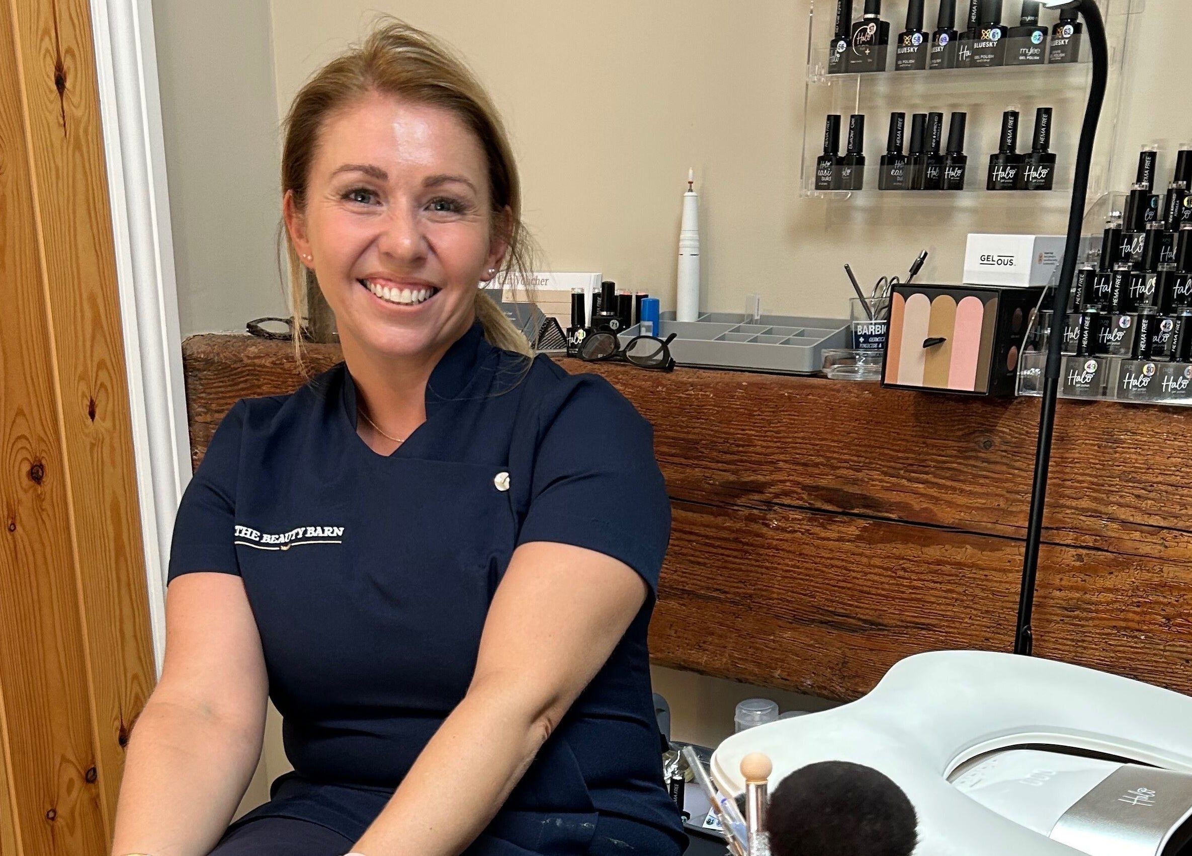 Smiling stylist at The Beauty Barn, Wilton, England, GB, surrounded by nail products.