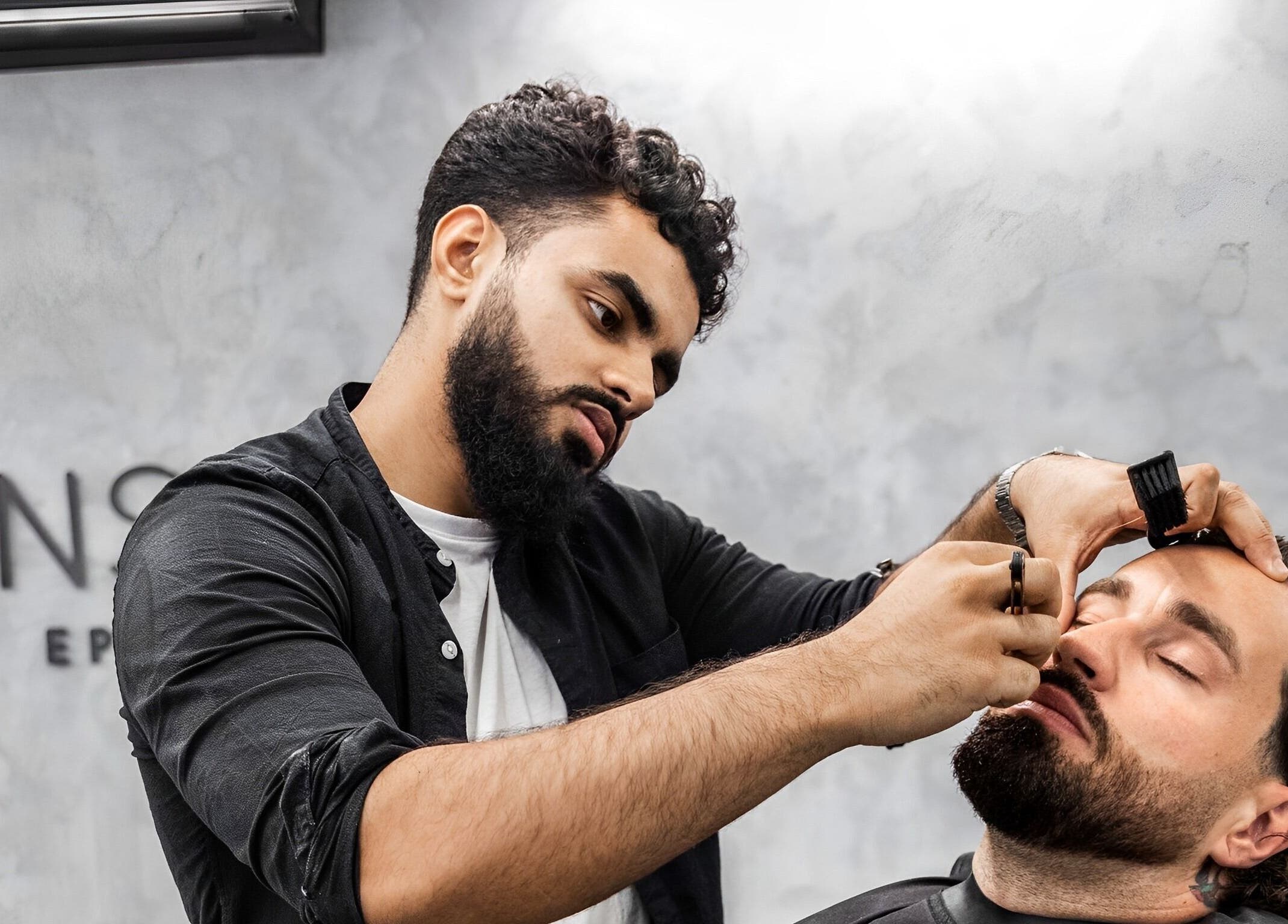 Professional grooming at Menspire Epsom, Epsom, England, GB. Barber shapes client's beard with precision tools.