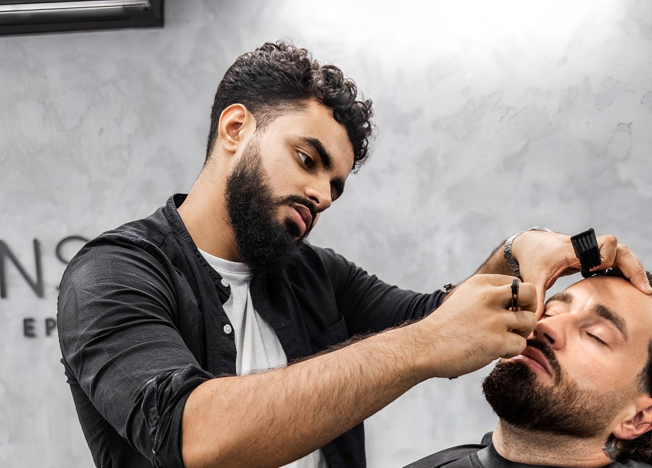 Professional grooming at Menspire Epsom, Epsom, England, GB. Barber shapes client's beard with precision tools.