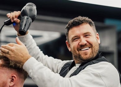 A barber at Lishmans Barbershop in Newcastle upon Tyne, England, styling a client's hair with a blow dryer.