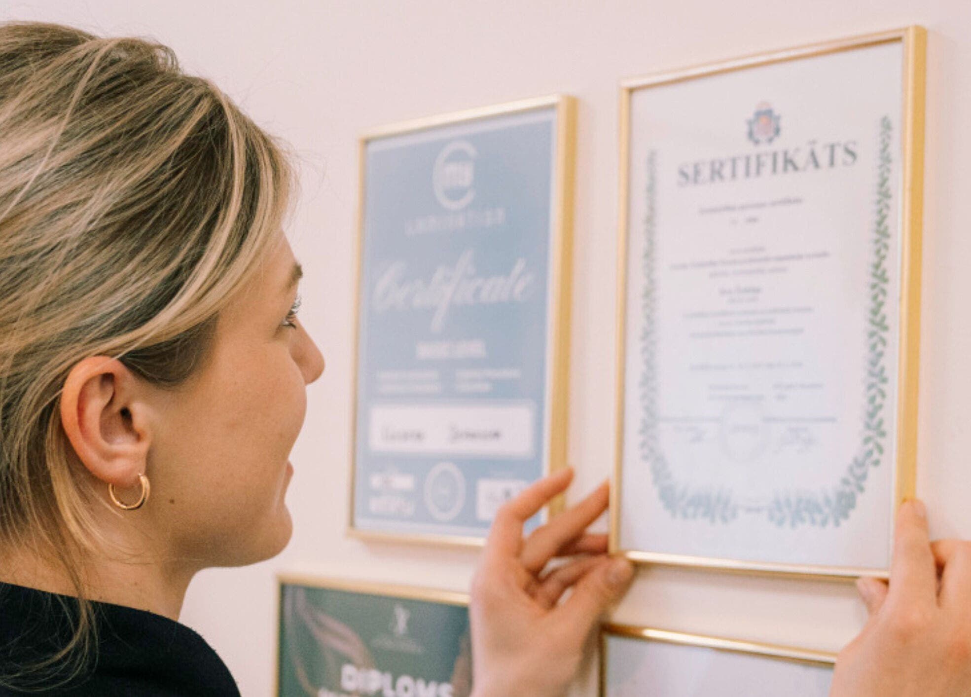 A staff member at FIZIO & BEAUTY HOUSE in Mārupe, Mārupes Novads, LV hangs certificates on a wall.