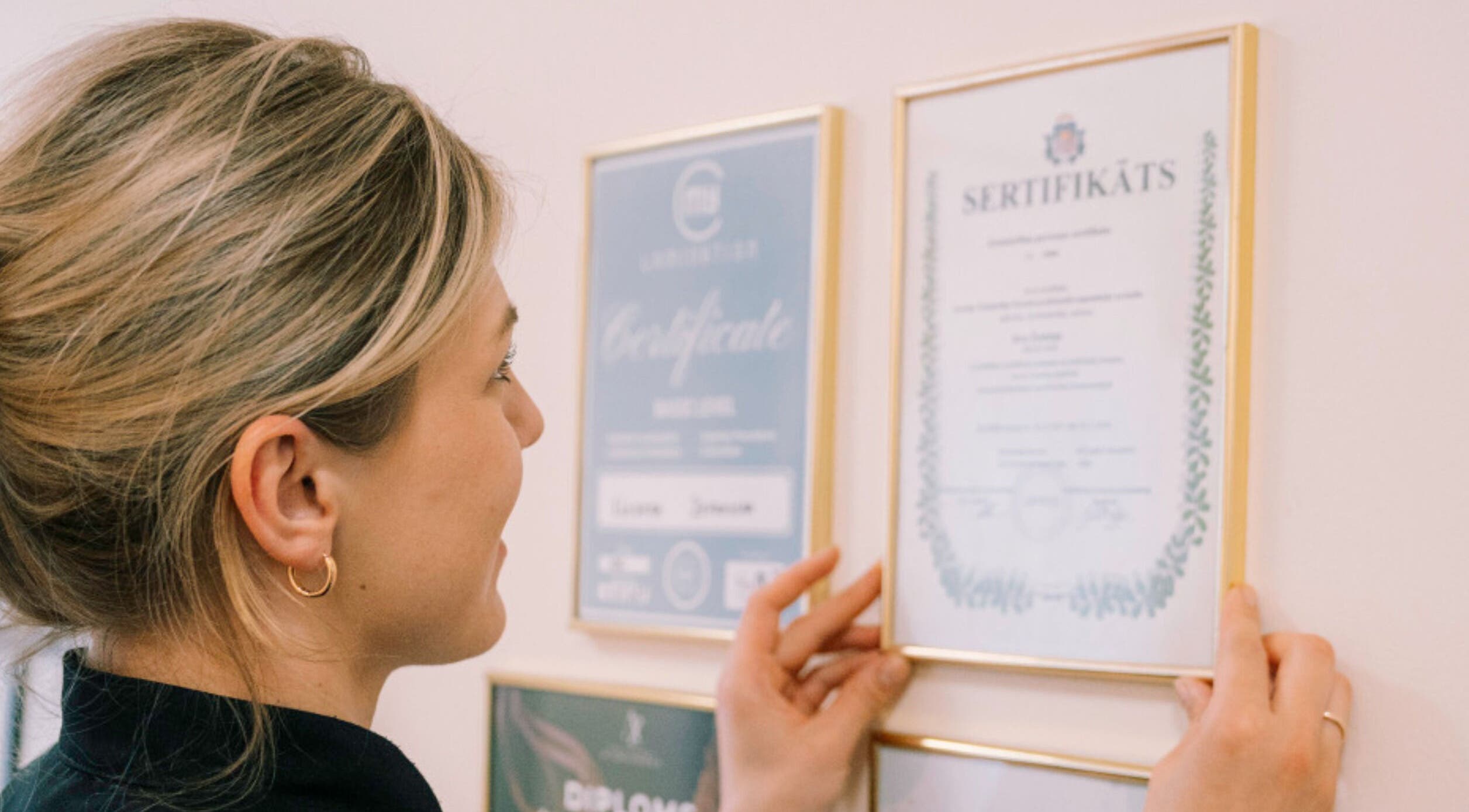 A staff member at FIZIO & BEAUTY HOUSE in Mārupe, Mārupes Novads, LV hangs certificates on a wall.