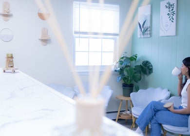 Woman sipping tea at Calm And Cozy Spa, Long Branch, New Jersey. Serene white decor with plants and art.