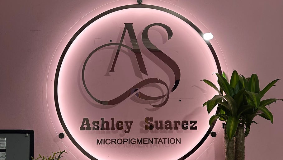 Logotipo iluminado de Ashley Suárez Micropigmentation en Guayaquil, Guayas, EC con fondo rosa y plantas verdes.