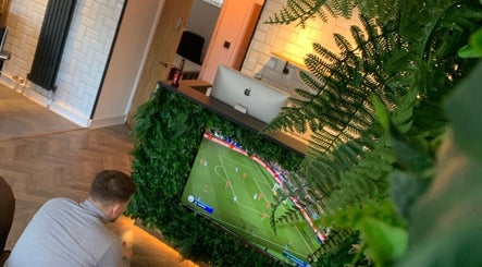 A stylish reception area at Cutthroat Jacks, Stockport, England, GB, featuring lush greenery and a TV showing a game.