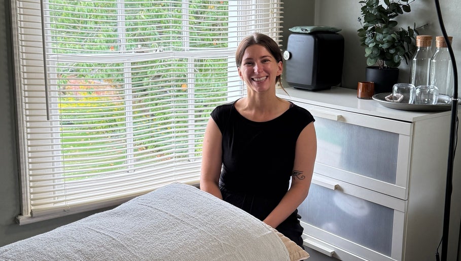 Smiling woman in therapy room at Root and Remedy Wellness, Purleigh, England, GB, beside a massage table.
