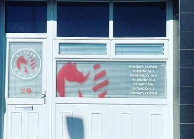 Exterior of The Barber Rhino in Carluke, Scotland, GB, featuring logo and opening hours on the glass door.