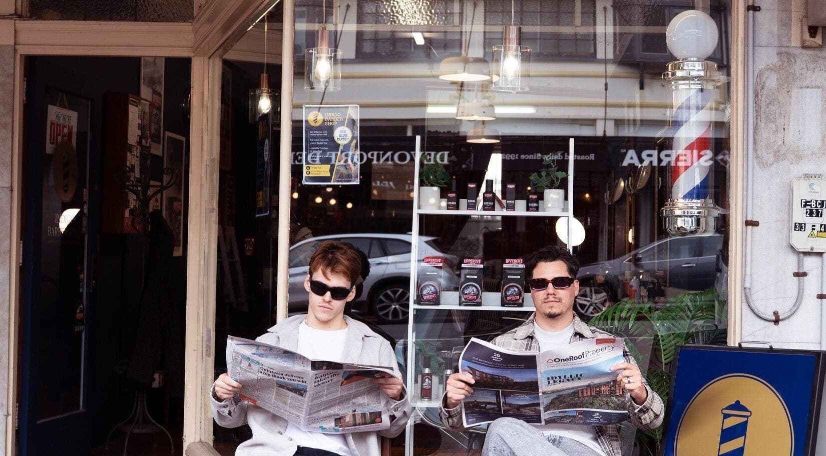Two customers reading newspapers outside Indigo Barbers, Auckland, Auckland, NZ, under classic barbershop pole.