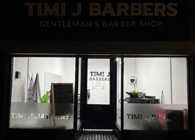 Entrance of TIMI J BARBERS in Paisley, Scotland, GB, warmly lit at night with welcoming decor.