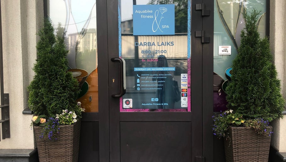 Entrance to Aquabike fitness & Spa in Rīga, LV, framed by plants, showcasing business hours and contact info.