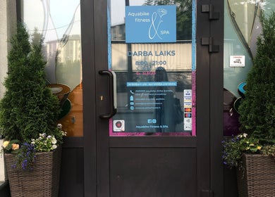 Entrance to Aquabike fitness & Spa in Rīga, LV, framed by plants, showcasing business hours and contact info.