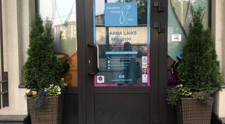 Entrance to Aquabike fitness & Spa in Rīga, LV, framed by plants, showcasing business hours and contact info.