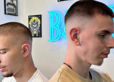 Δύο άνδρες με κούρεμα στη Blade Barbershop Skg, Ευκαρπία, GR δίπλα σε φωτεινή επιγραφή.
