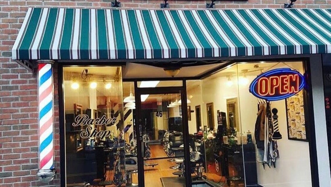 Front view of Luisthebarber-evolutionpatchogue in Patchogue, New York, US with striped awning and neon open sign.