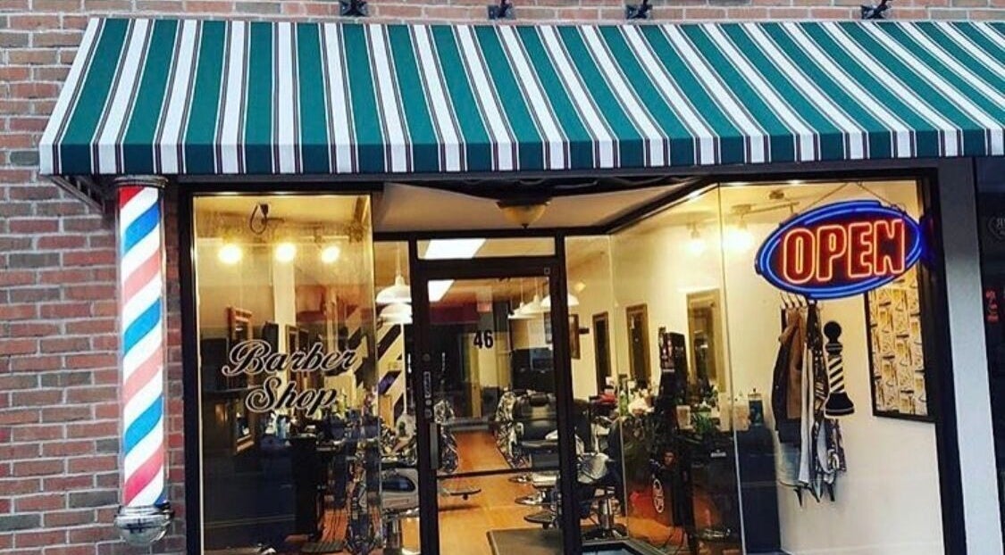 Front view of Luisthebarber-evolutionpatchogue in Patchogue, New York, US with striped awning and neon open sign.