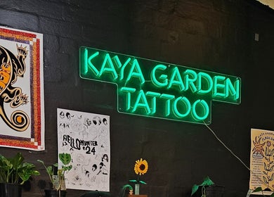 Neon sign at Kaya Garden Tattoo Haymarket, NSW, AU, surrounded by colorful tattoo posters and plants.