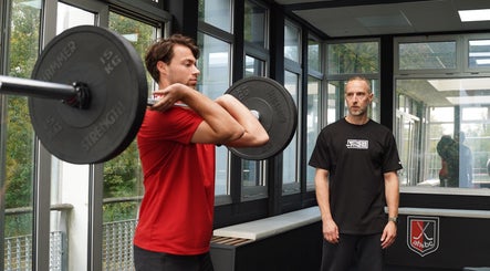 Man lifts barbell under trainer's guidance at WITNESS THE FITNESS at AH&BC PERFORMANCE GYM, Amstelveen, Noord-holland, NL.