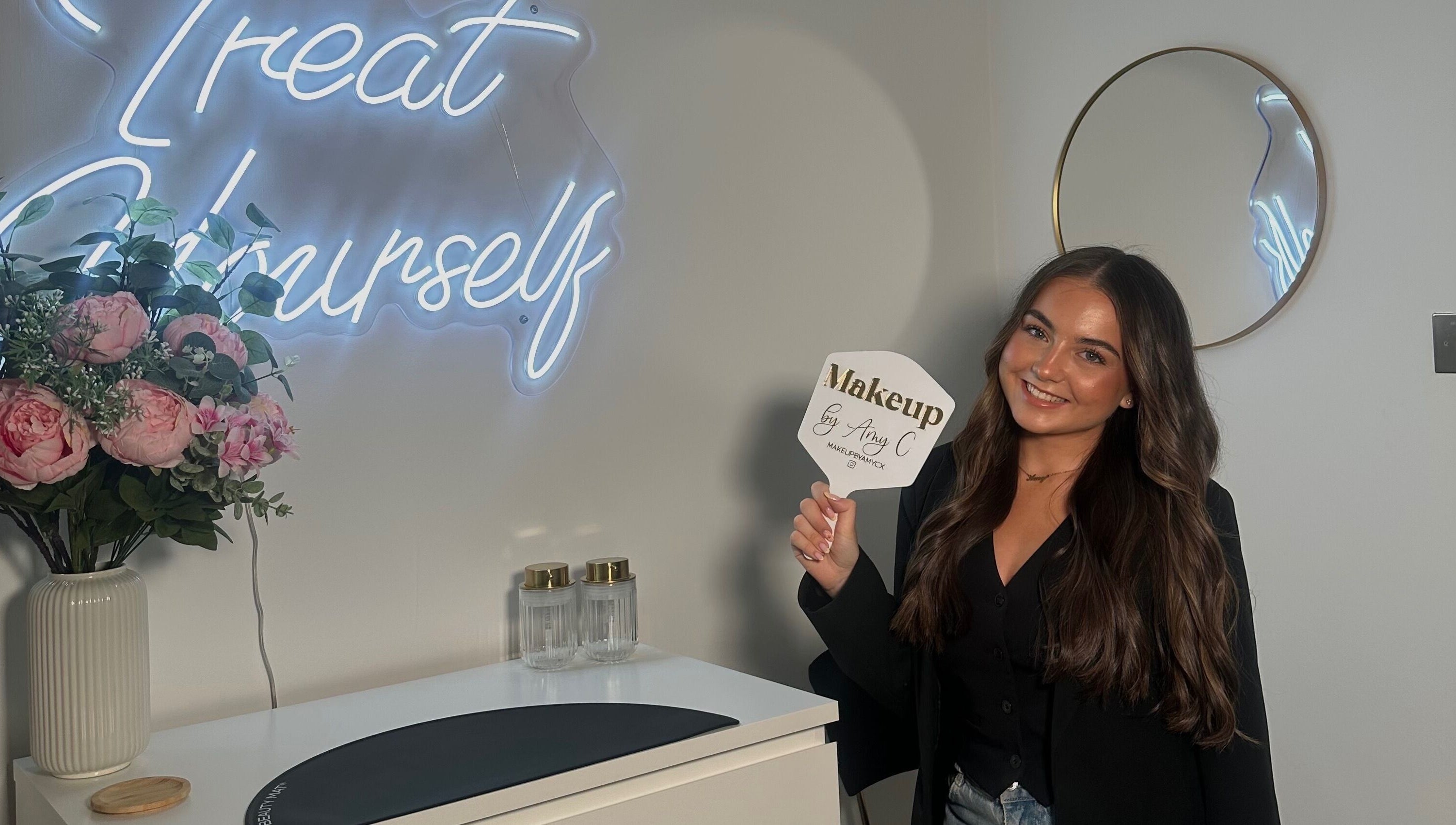 Inside MakeupbyAmyC in East Kilbride, Scotland; woman holds branded mirror, elegant decor visible.