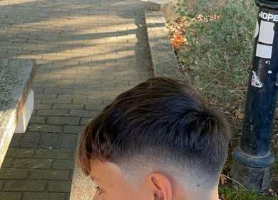 Corte moderno de cabelo curto em jovem na G Barbershop, Alcabideche, Lisboa, PT, com fundo de rua arborizada.
