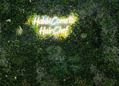 Neon sign saying 'Habibi, Come to HairChef' on lush greenery at Hairchef & Dociä Salon LLC, Dubai, AE.