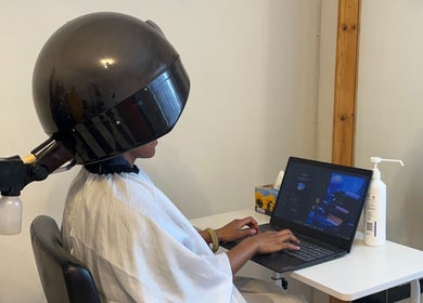 Client under hair dryer working on laptop at Braidsbyvuyo - Natural Hair Salon, Sandton, Gauteng, ZA.