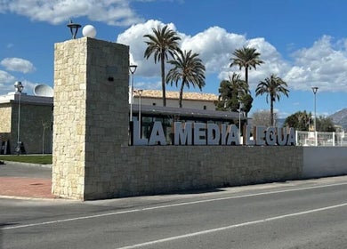 LaMediaLeguaHair entrance in Comunidad Valenciana, ES with palm trees and stone structure under a blue sky.