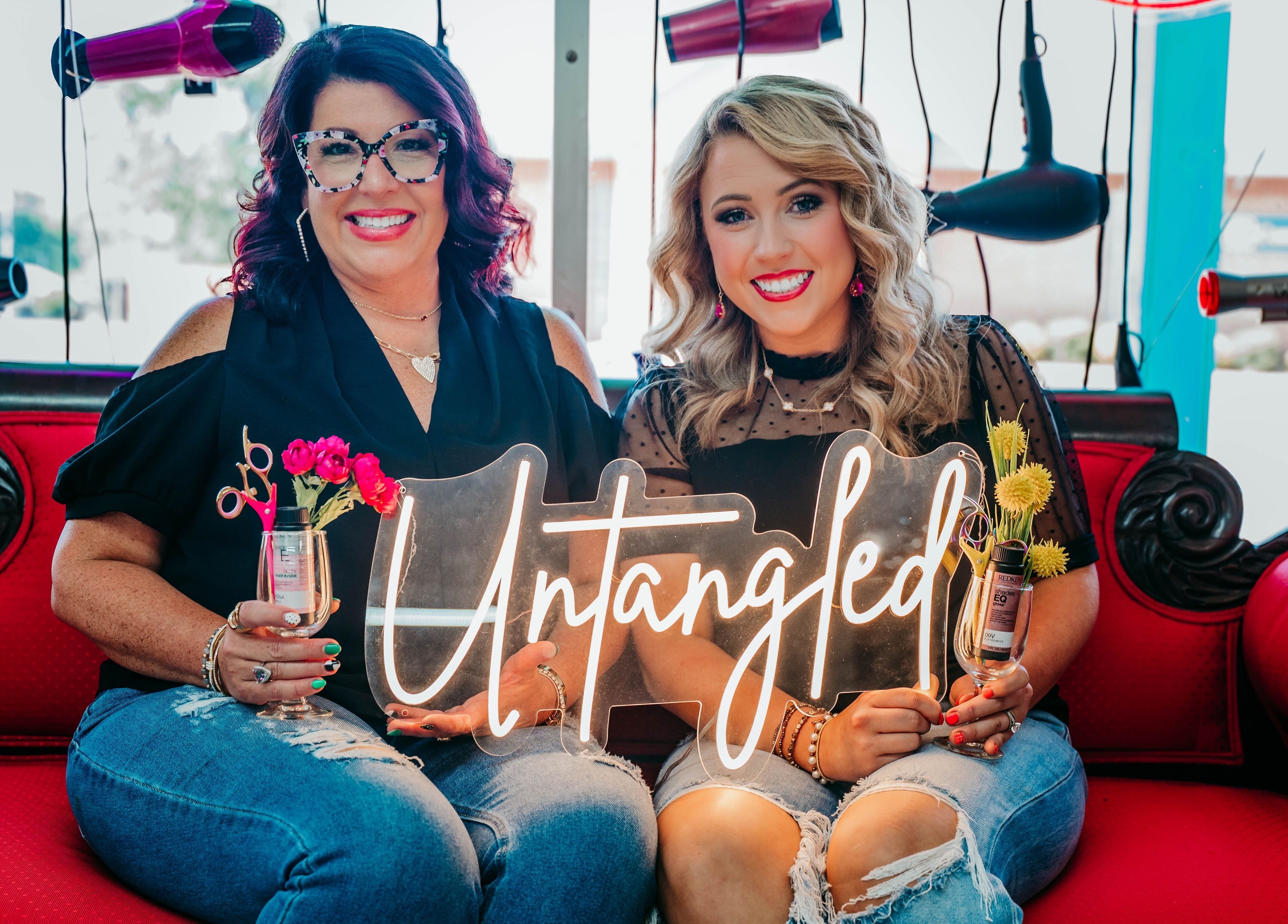 Stylists at Untangled Salon in Hampton, Arkansas, smiling on a red sofa, surrounded by hanging hairdryers.