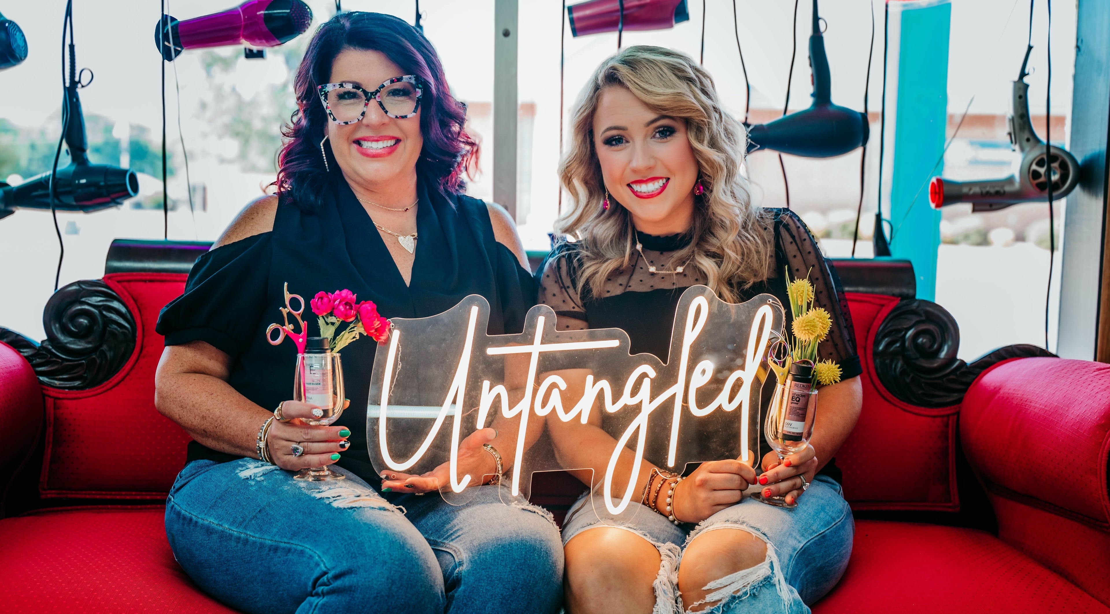 Stylists at Untangled Salon in Hampton, Arkansas, smiling on a red sofa, surrounded by hanging hairdryers.