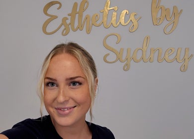 Smiling individual at Esthetics by Sydney in South Glengarry, Ontario, CA with logo in the background.
