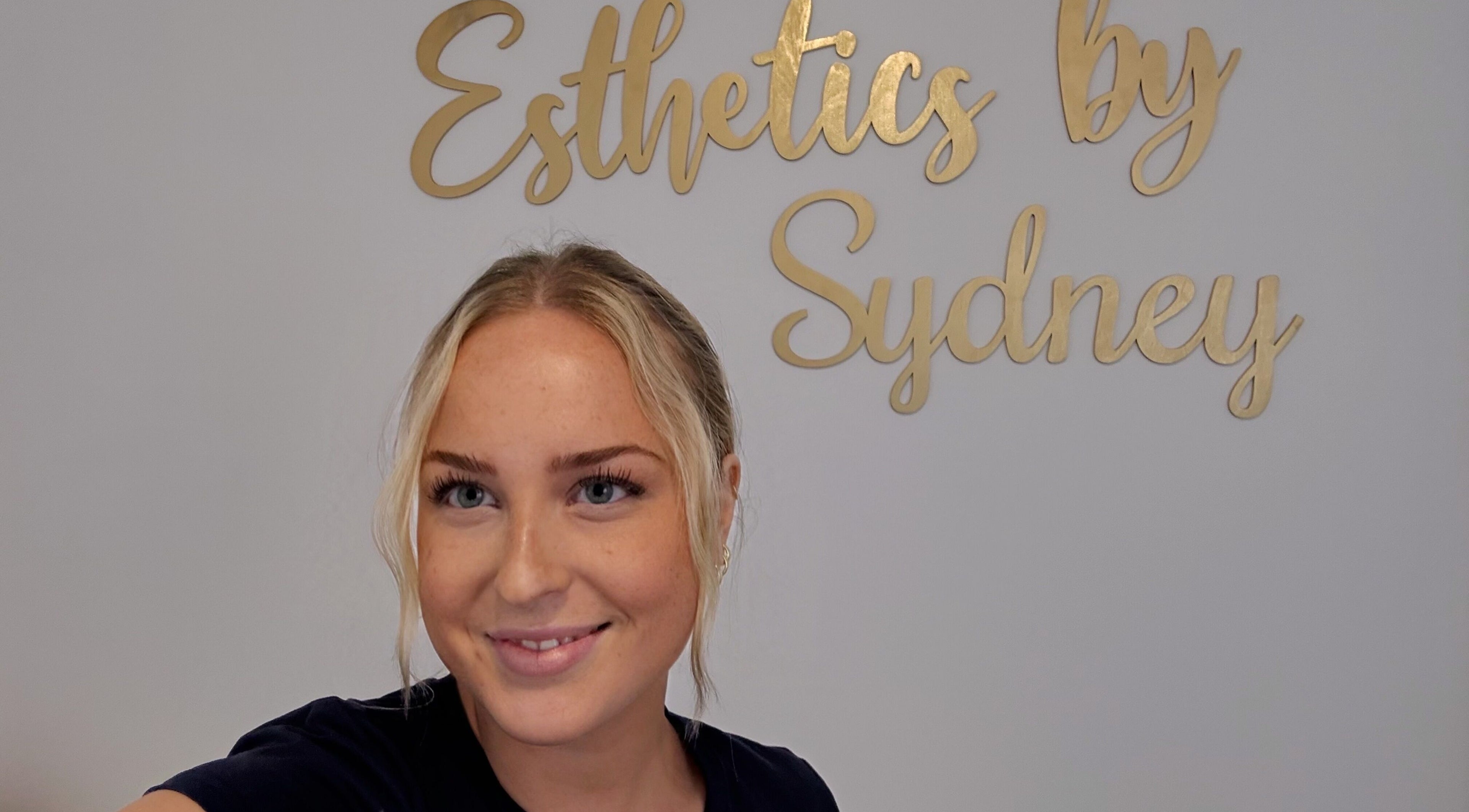 Smiling individual at Esthetics by Sydney in South Glengarry, Ontario, CA with logo in the background.