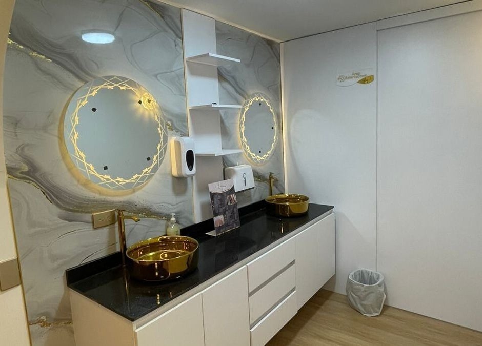 Baño moderno en Emy Study Nails, Bogotá, Bogotá, CO con espejo iluminado y lavamanos dorados.