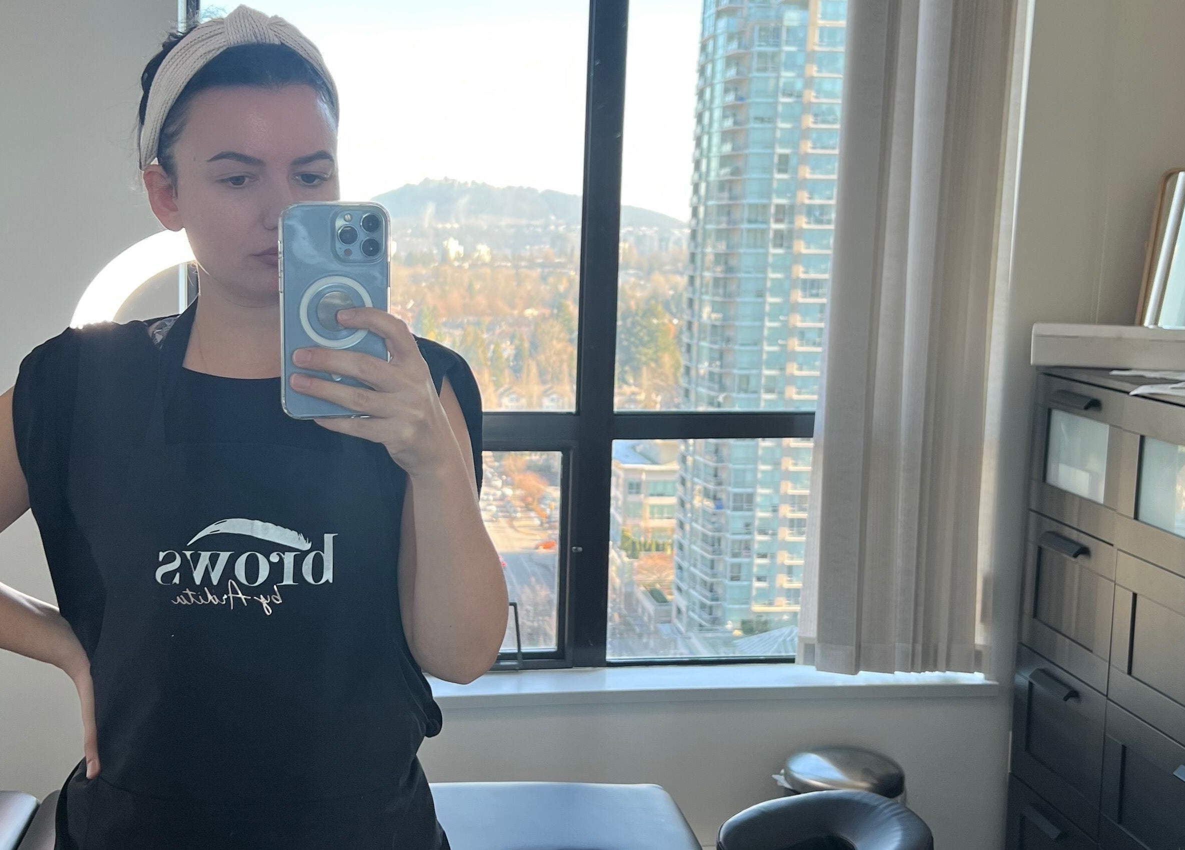 Professional at Brows by Ardita in Coquitlam, BC, captures a studio selfie with cityscape backdrop.