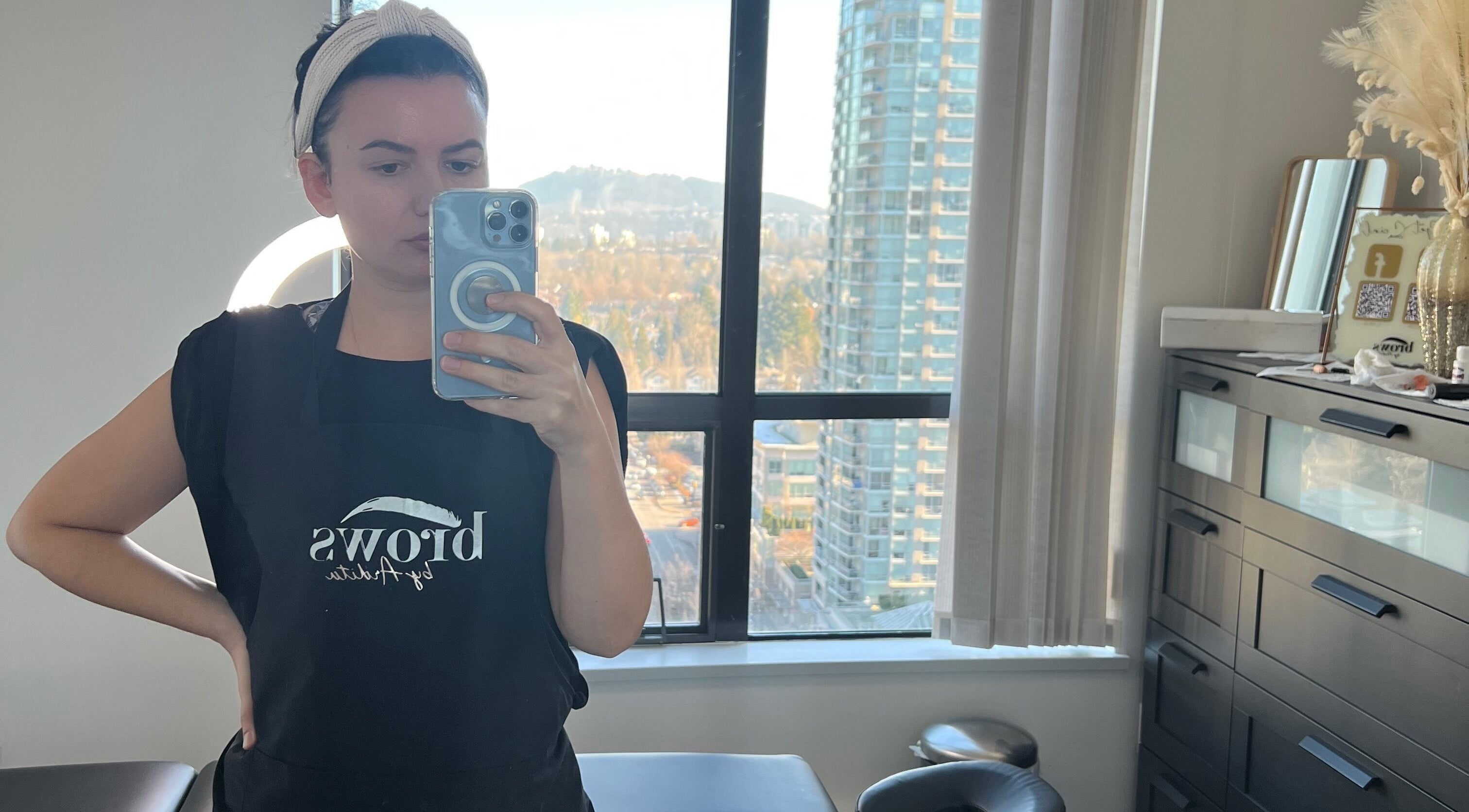 Professional at Brows by Ardita in Coquitlam, BC, captures a studio selfie with cityscape backdrop.