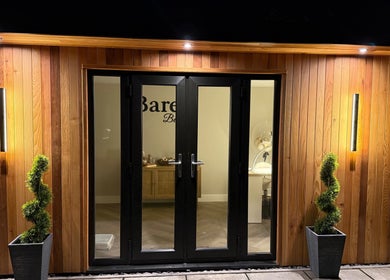 BARE Beauty entrance at night, Birstall, England, GB. Wooden doors with soft lighting and greenery.