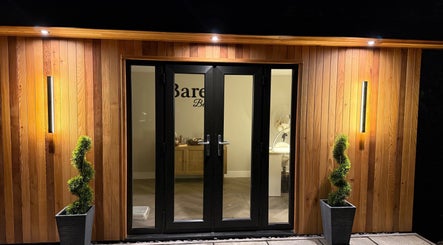 BARE Beauty entrance at night, Birstall, England, GB. Wooden doors with soft lighting and greenery.