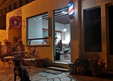 Η είσοδος του Xonikis Men’s Hair Studio στη Μεγάλη Παναγία, GR, φωτισμένη τη νύχτα με καλαίσθητες λεπτομέρειες.
