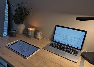 Cozy workspace at Silk Touch Tattoos, Colchester, England with a laptop, tablet, and decor on wooden desk.