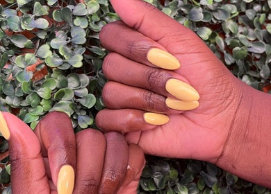 Stylish yellow manicure on dark skin at PS Nail Bar, Randburg, Gauteng, ZA, accentuated by green foliage.