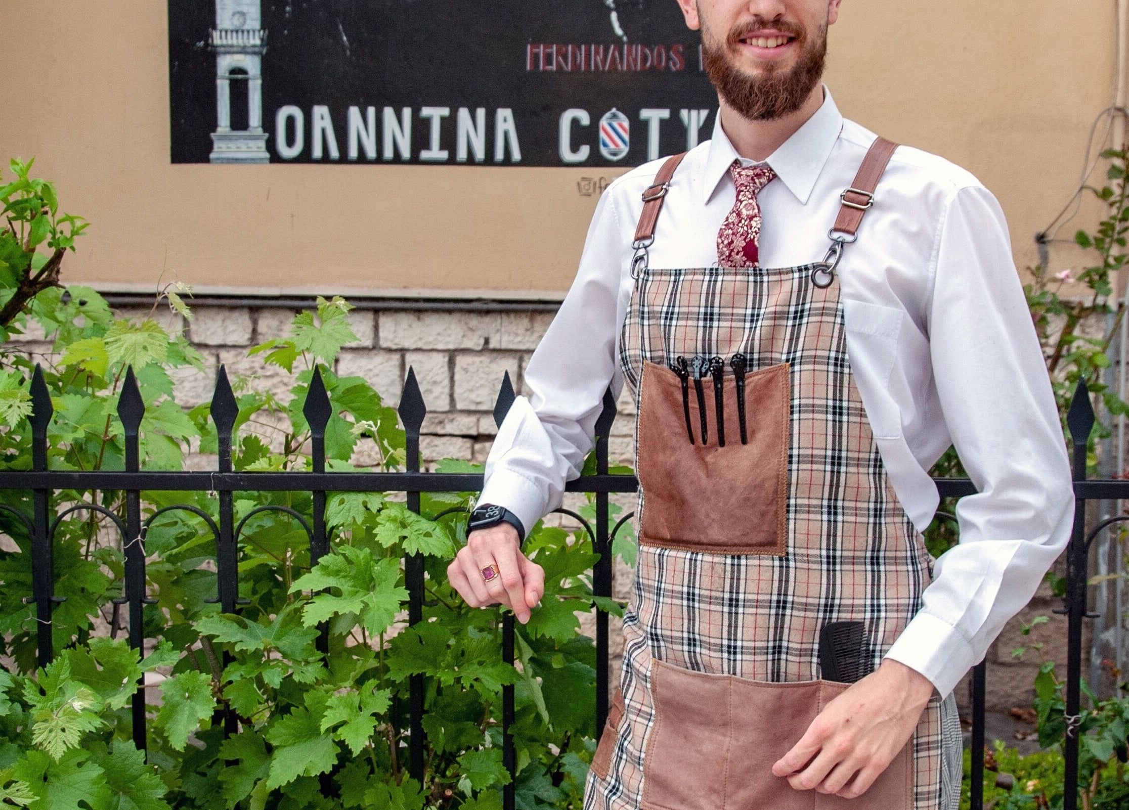 Έξω από το Ferdinandos Barbershop, Ιωάννινα, GR, ένας κουρέας με καρό ποδιά και κομψή εμφάνιση.