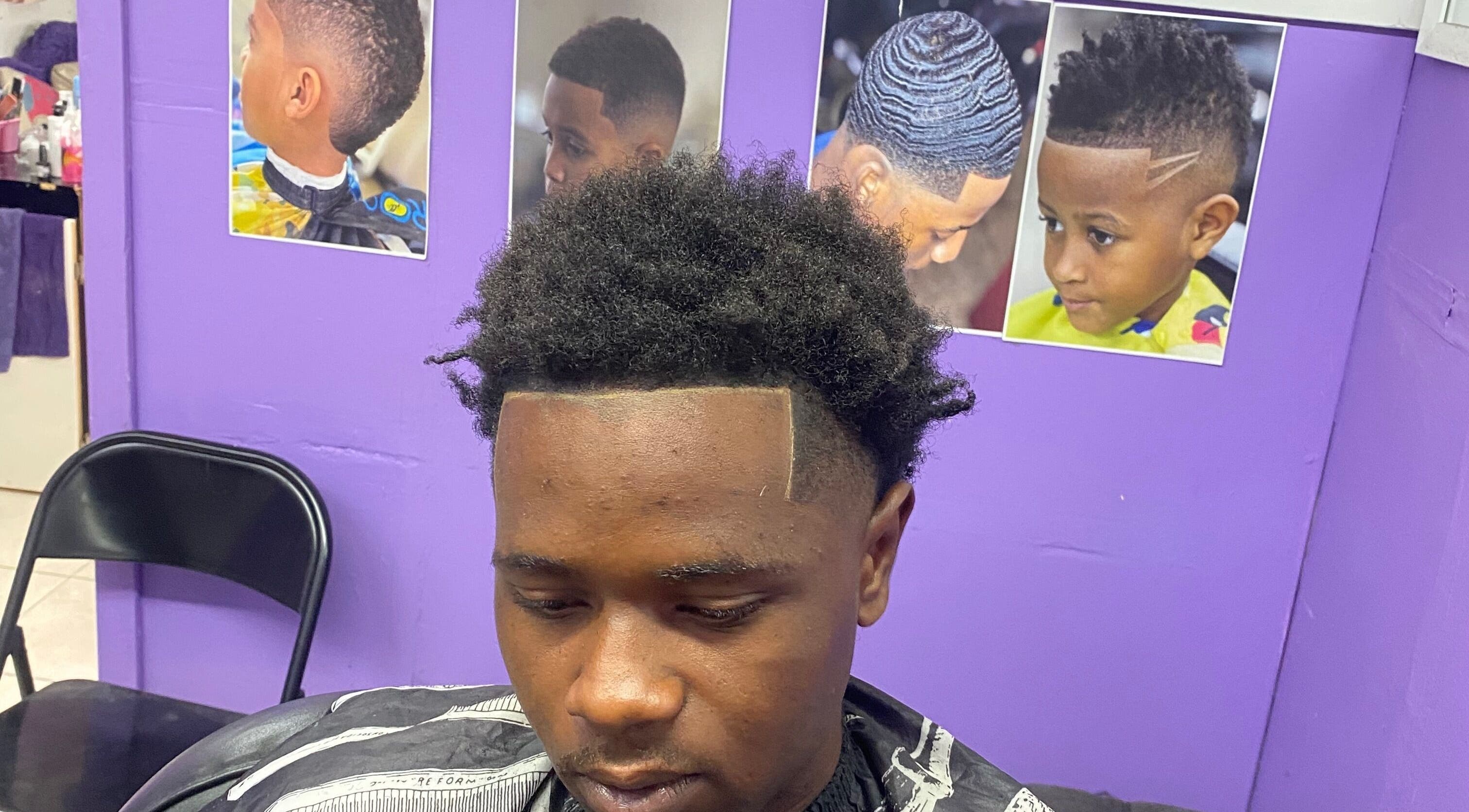 Man with stylish haircut at Dre Cool Cutz, Nassau, New Providence, BS, seated in front of hair style posters.