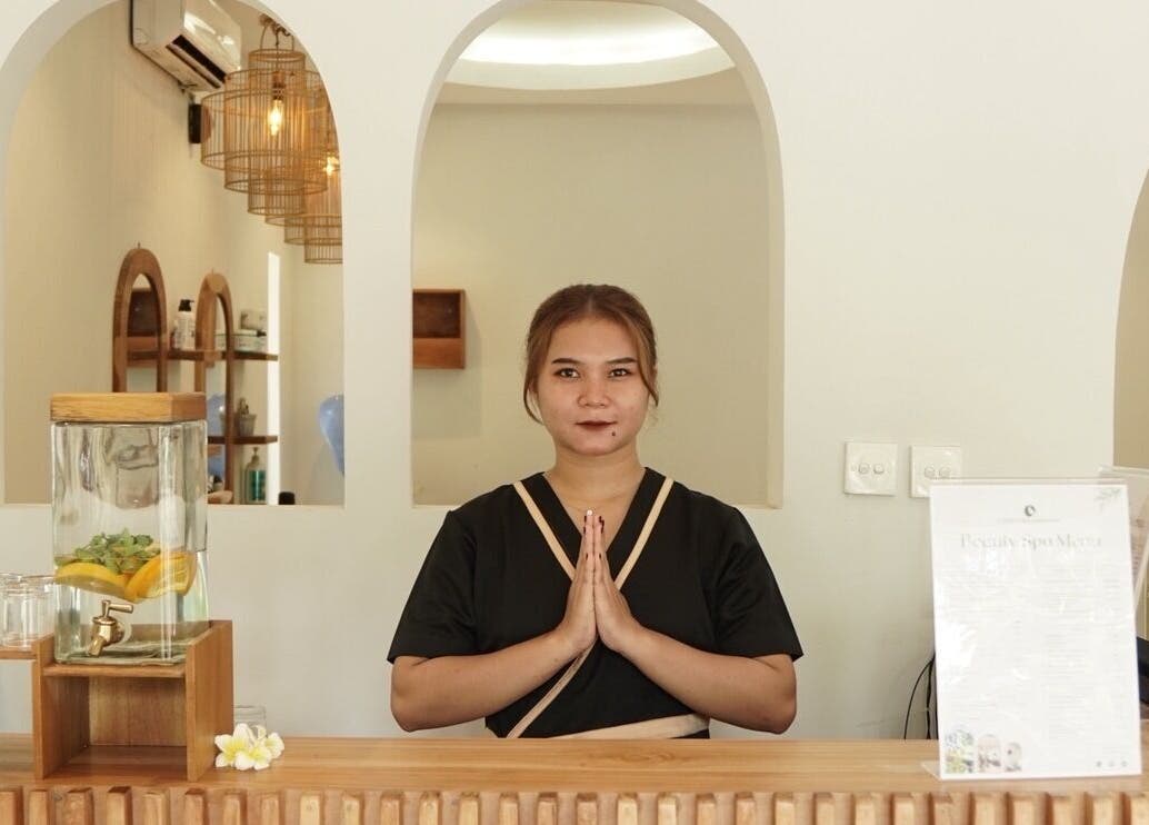 Receptionist welcoming guests at KU Salon & Spa, Nusa Tenggara Barat, ID with a tranquil smile and clasped hands.