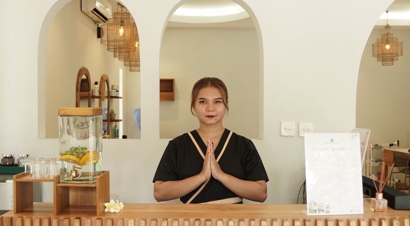 Receptionist welcoming guests at KU Salon & Spa, Nusa Tenggara Barat, ID with a tranquil smile and clasped hands.