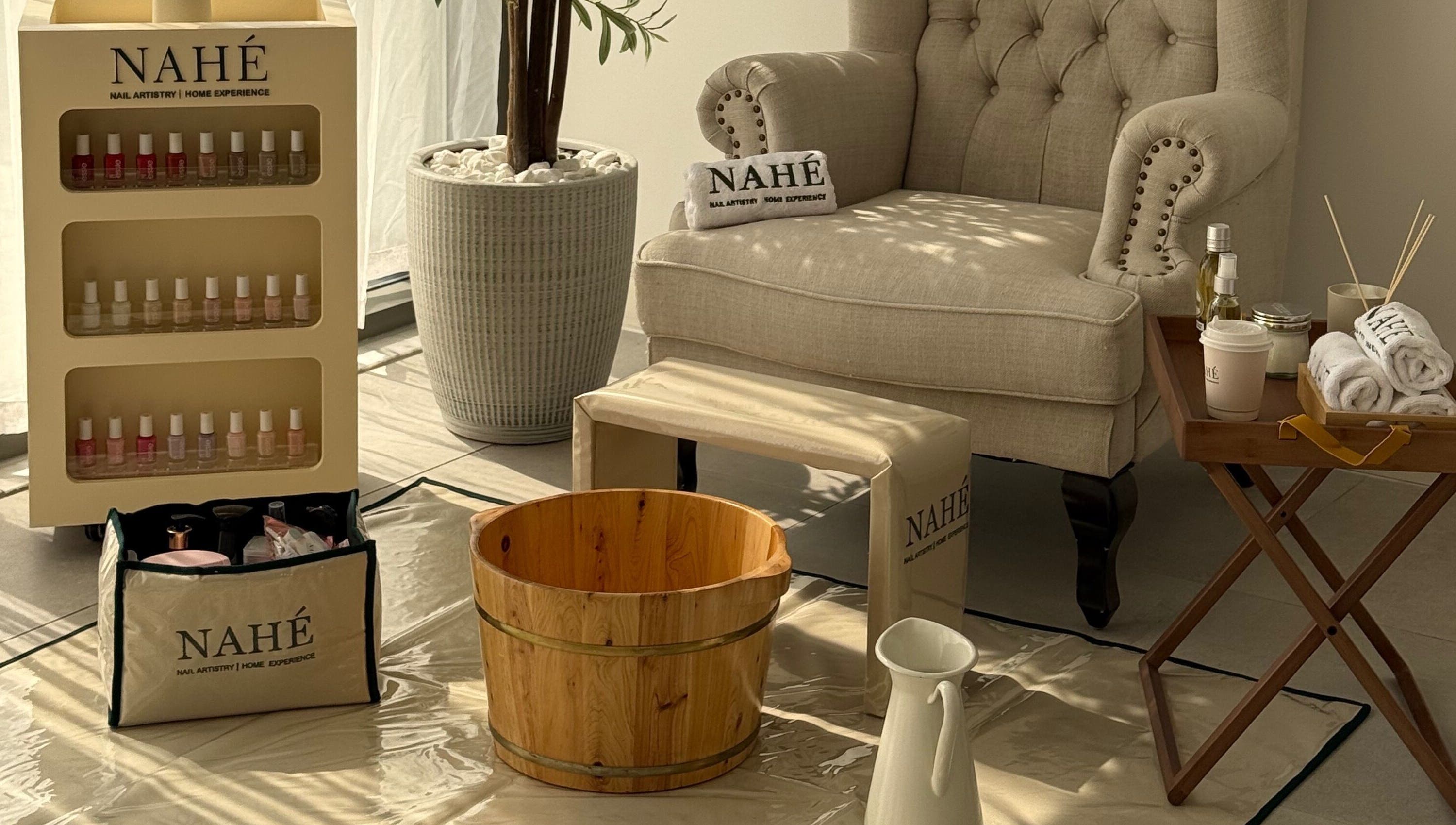 Elegant spa setup at Nahé Beauty Home Service Salon, Dubai, AE with armchair, wooden tub, and beauty products.
