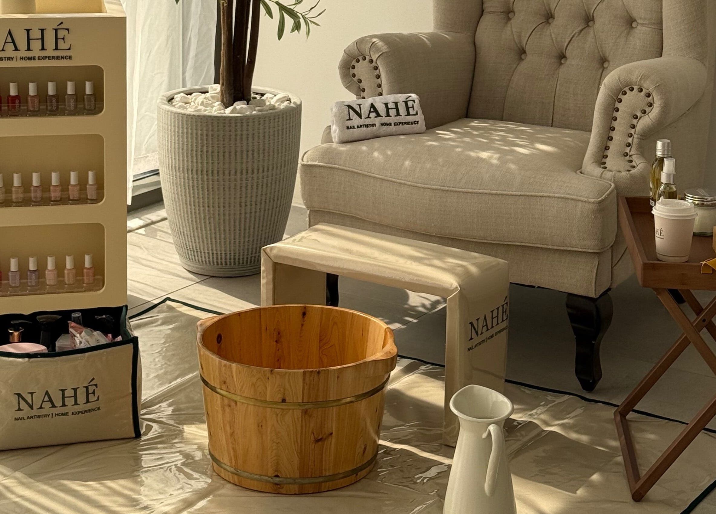 Elegant spa setup at Nahé Beauty Home Service Salon, Dubai, AE with armchair, wooden tub, and beauty products.