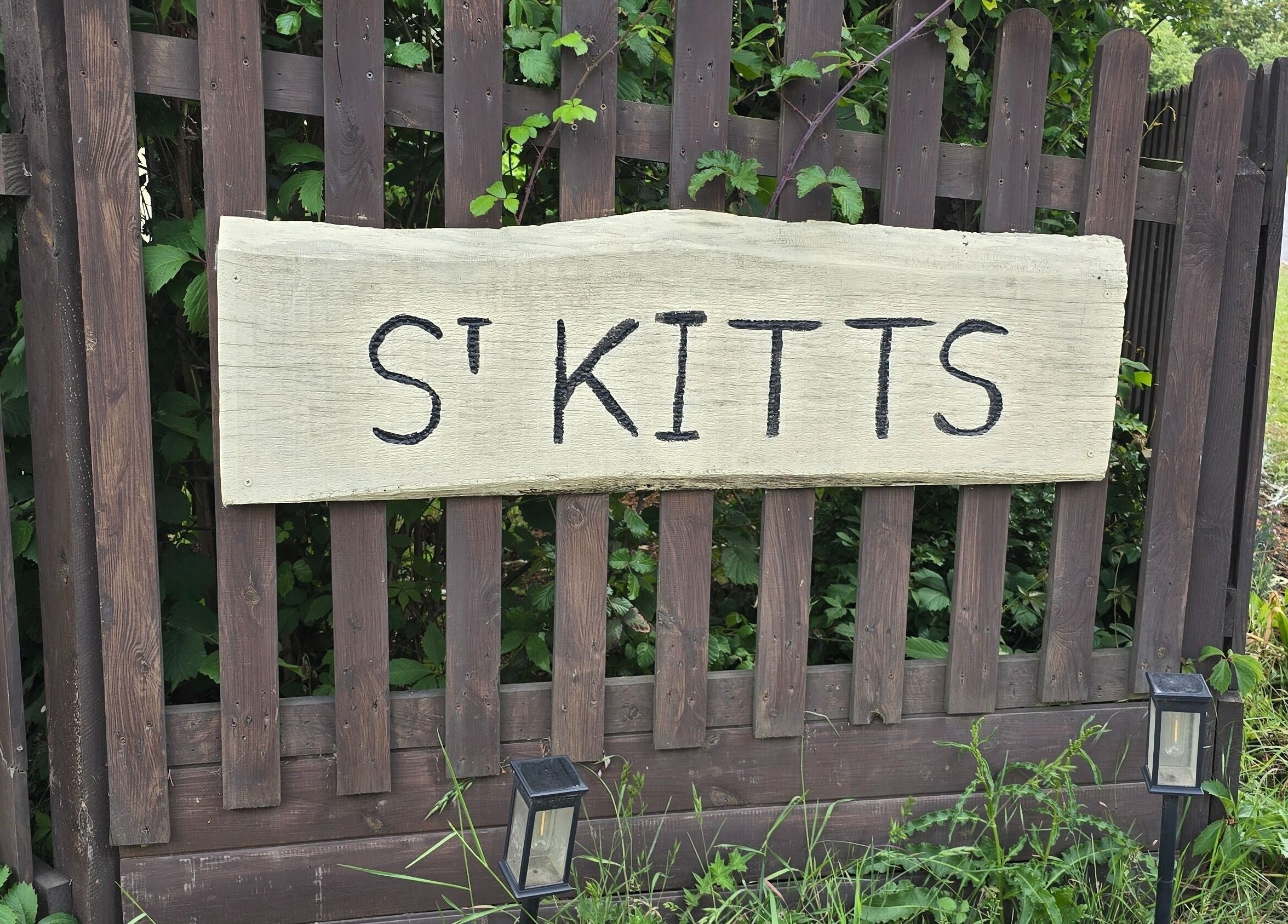 Wooden sign by The Cabin, Black Notley, England, GB with 'St Kitts' text surrounded by greenery.