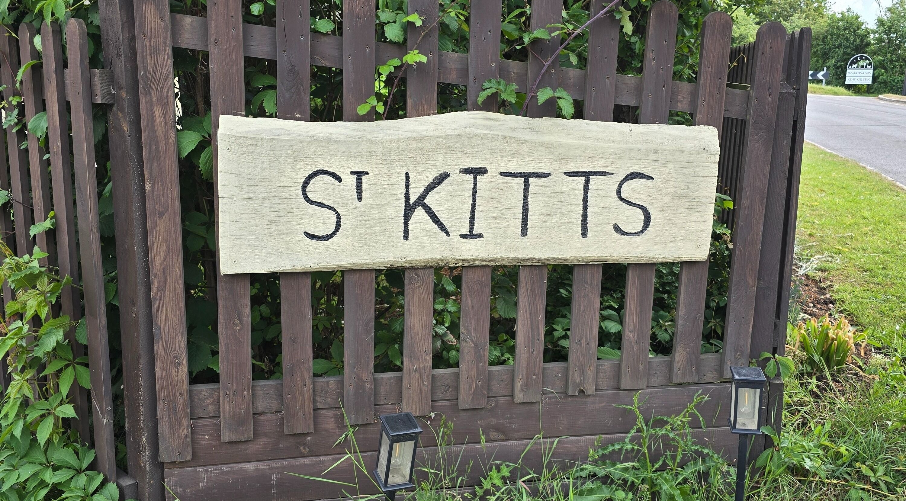 Wooden sign by The Cabin, Black Notley, England, GB with 'St Kitts' text surrounded by greenery.
