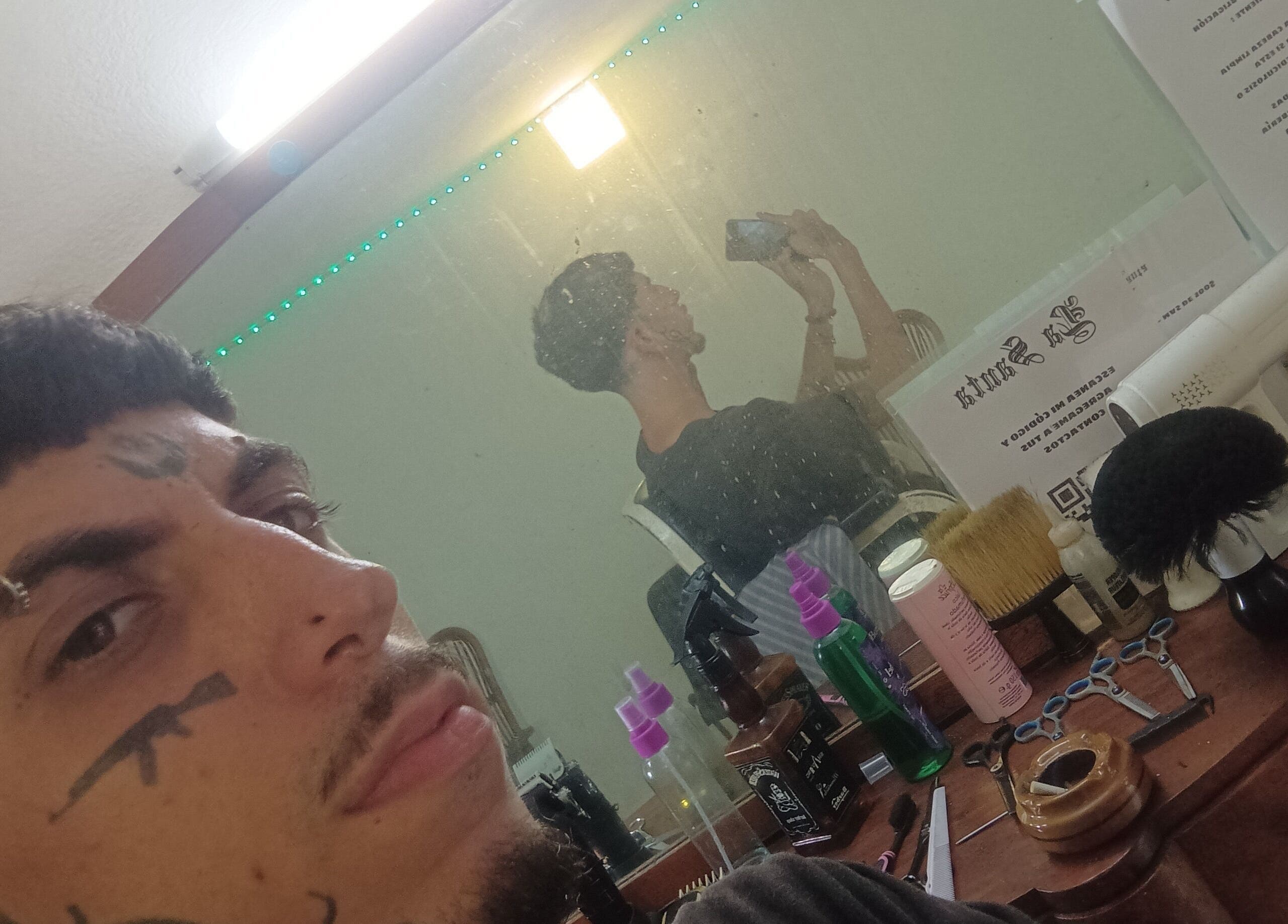 Estilista en La Santa Barbería, La Habana, rodeado de herramientas de barbería y un espejo iluminado.