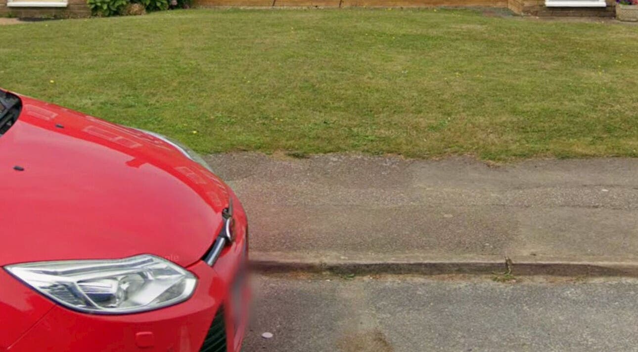 A vibrant red car parked outside NailsbyNays in Hitchin, England, GB, in front of a lush green lawn.