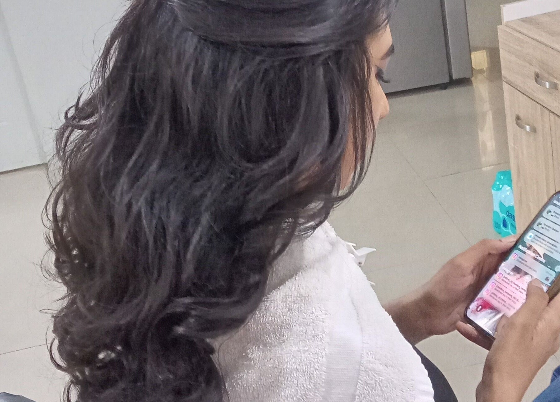 Client with styled hair browsing phone at R. Luxe Hair Studio, Chaguanas, Chaguanas, TT.